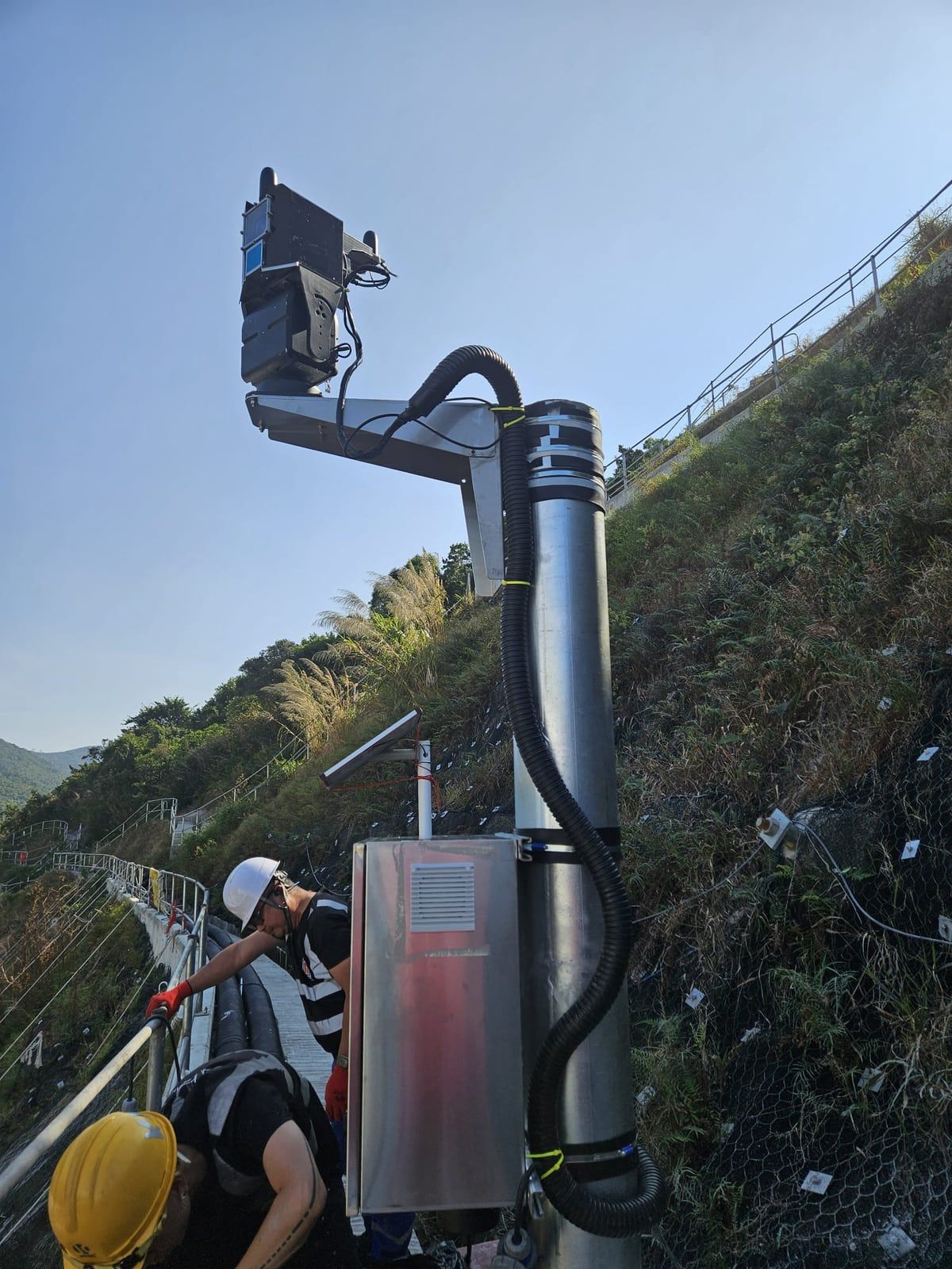 A Kodifly LiDAR unit and equipment cabinet on a pole above a protected hillside, with technicians at work